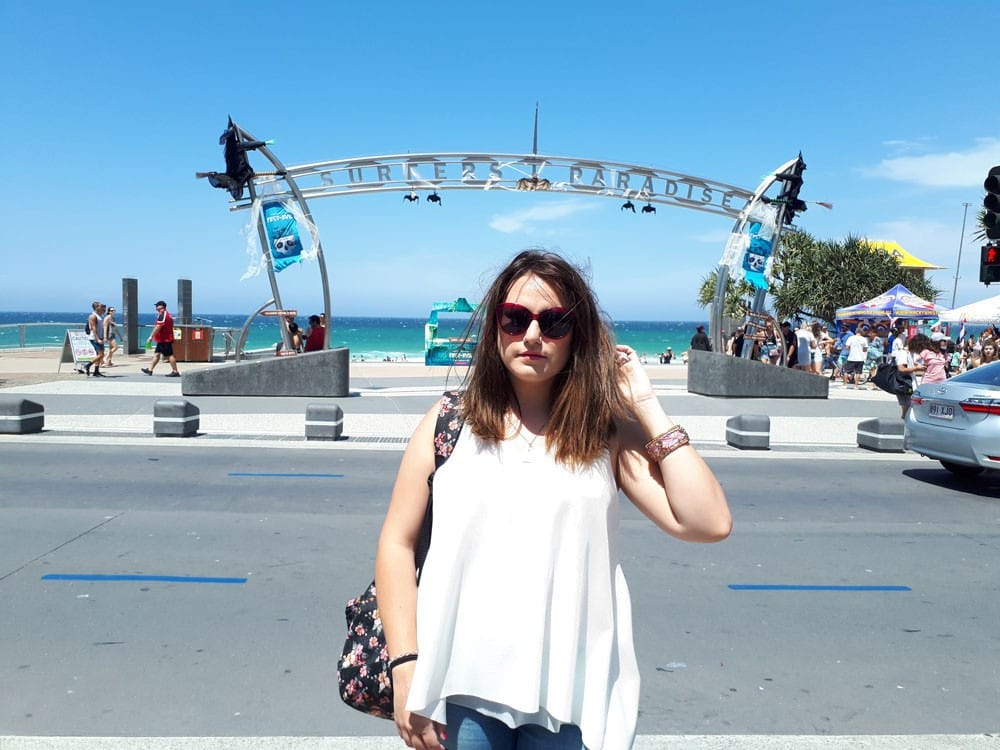 chica morena con gafas de sol y una camiseta blanca posando a cámara delante del letrero de "Surfers Paradise" de Gold Coast